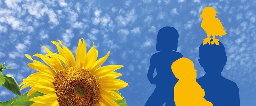 A sunflower against a blue sky with small clouds and the silhouettes of three people in the background