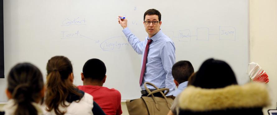 A photo of a male in a blue shirt and red tie looks at a class while standing at a white board