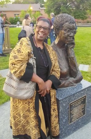 Photo of a smiling Sherry Williams in a black shirt and yellow and black patterned jacket standing next to the Gwendolyn Brooks statue in Bronzeville