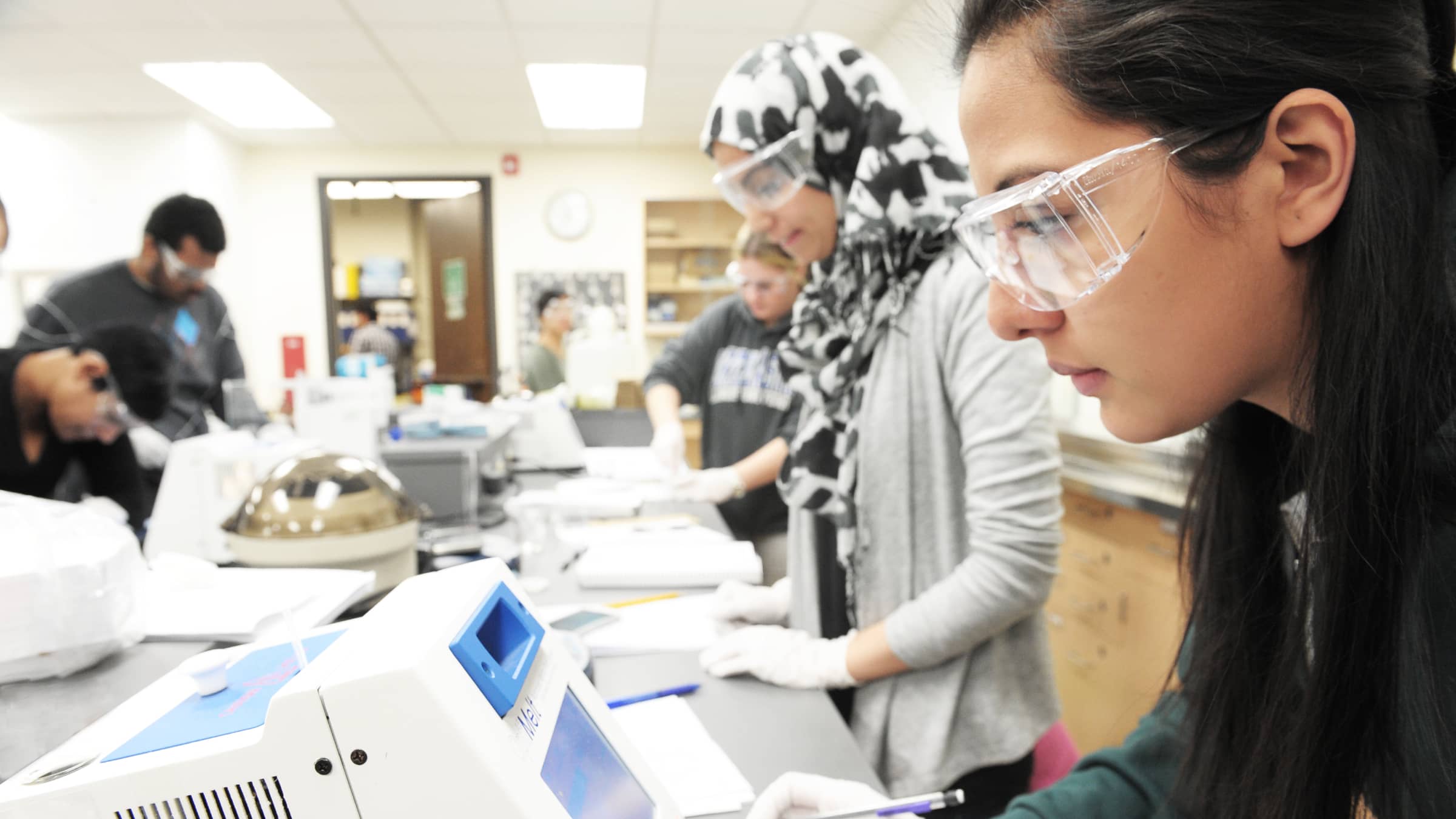 Female chemistry students wearing safety goggles analyze samples in a lab at NEIU