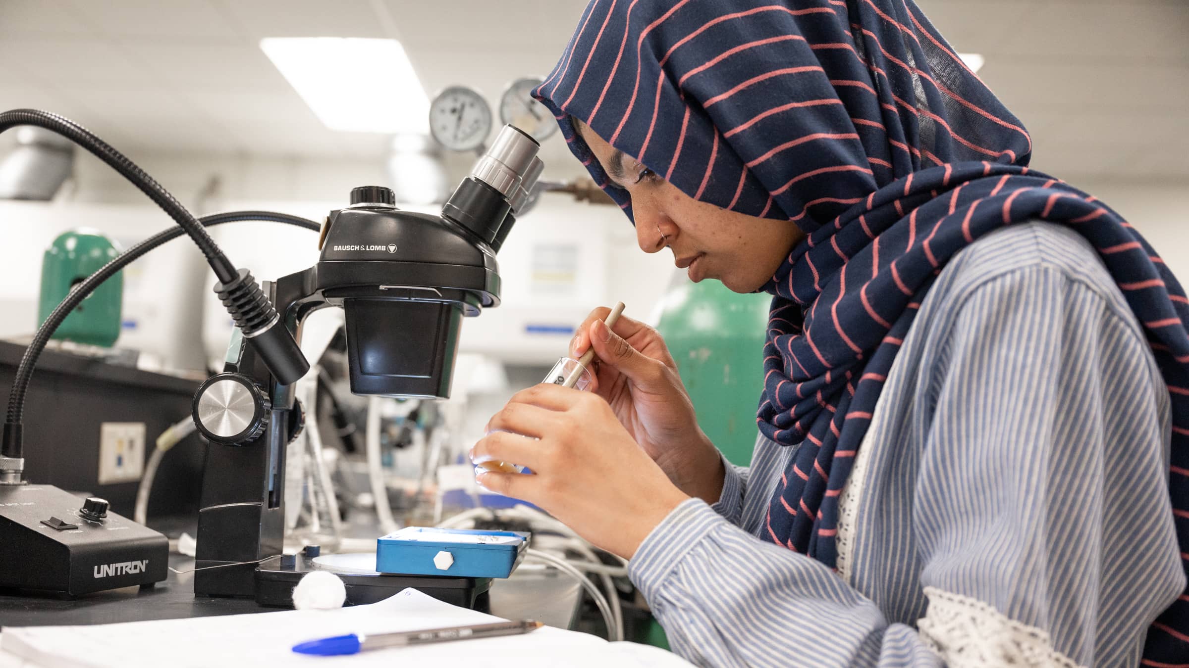 A female biology student wearing a hijab analyzes a sample in a test tube in front of a microscope in a lab at NEIU