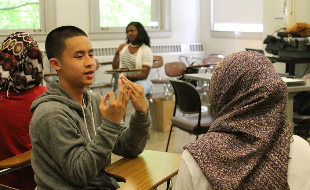 A male student facing a female student. The male student is using Sign Language. Both students are in a classroom.