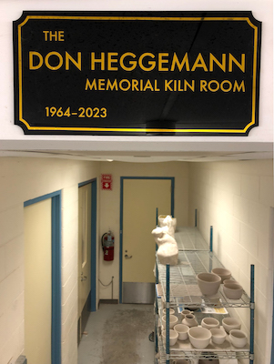 A photo of the Ceramics kiln room sign, which reads "The Don Heggemann Memorial Kiln Room 1964-2023" in gold letters on a black background, hanging above a rack with various ceramics. 