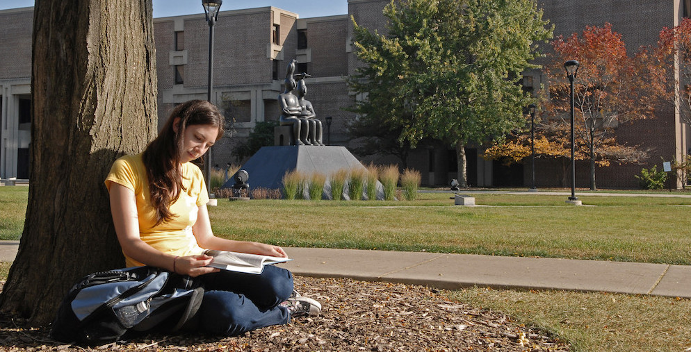 A student sits by a tree reading a book. 