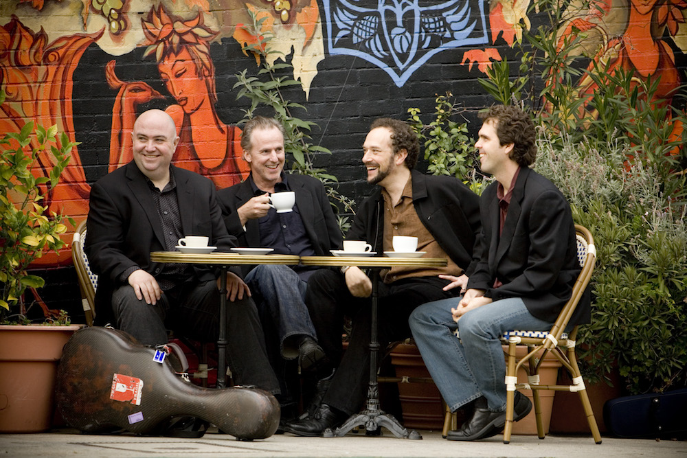 The four male members of the Los Angeles Guitar Quartet sitting at a table, drinking coffee and smiling with a colorful mural and plants in the background