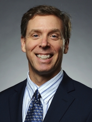 Photo of Thomas Philion in a dark suit, blue and white striped shirt and tie, smiling in front of a gray background. 