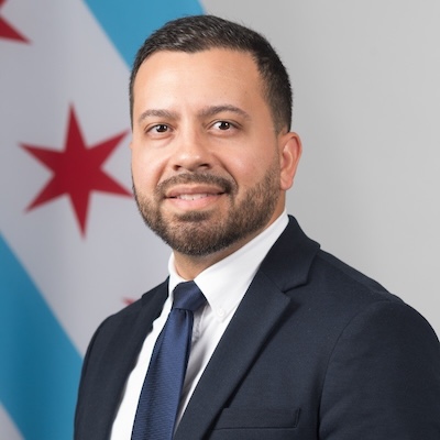 A headshot photo of Daniel Correa Jr. wearing a dark colored suit with white collard shirt and dark blue tie against a gray background with a partial image of the Chicago flag.