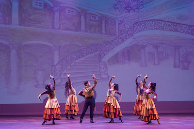 Six costumed dancers on stage raise their arms in unison