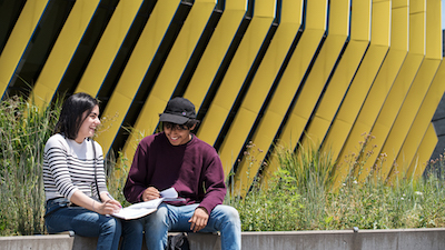 Two students sit on a bench outside of the El Centro building