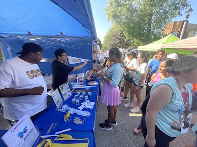 A photo of James Taylor (right) and Muhammad Elbadawi (left) working the Northeastern booth at the Corrida del Mariachi. Elbadawi is holding a CCICS pennant, showing an attendee what they won on the prize wheel. 