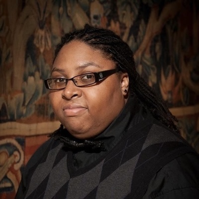 A headshot photo of Jerusalem L. Singleton wearing a black and gray sweater vest and black shirt, bowtie and glasses. The background is a muted-colored floral pattern.