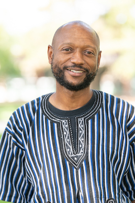 Photo of Kamau Rashid smiling in a black, white and blue striped shirt.