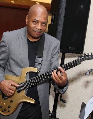 Keith Henderson wearing a gray jacket and black shirt holding a gold-colored guitar