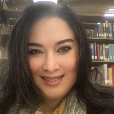 A headshot photo of Mayra Montante, smiling, wearing a yellow scarf and black jacket. Bookshelves are visible in the background.