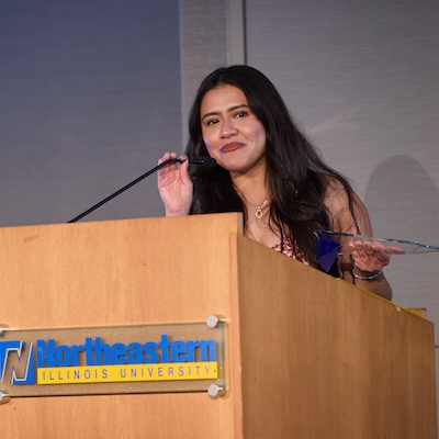 A photo of Aidé Hernández, Ph.D., speaking at a podium, accepting her Alumni Award.