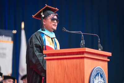 A photo of Alvin Gutierrez, wearing black graduation regalia and sunglasses, speaking at the Commencement podium. 
