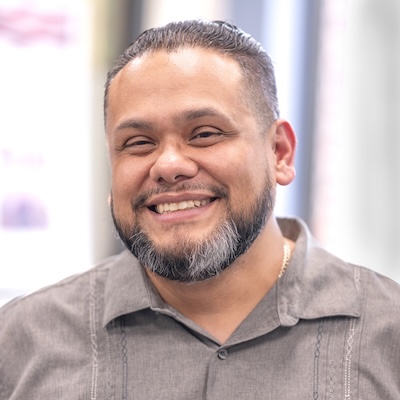 A headshot photo of Rogelio Villa Jr., smiling, wearing a gray collared shirt.