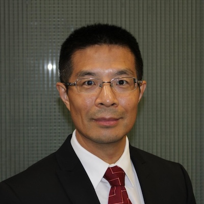 A headshot photo of Sheng Liu, Ph.D., wearing glassed and a dark suit with a white collared shirt and red tie, against a gray tiled background.