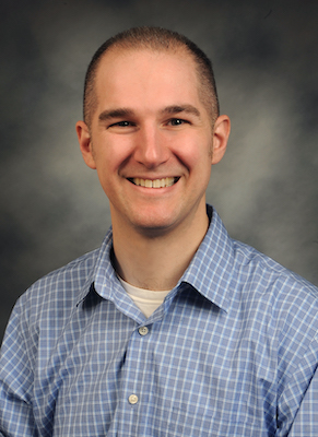 Photo of Aaron Schirmer smiling, wearing a blue and white checkered shirt against a gray background