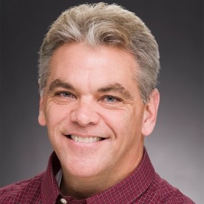 A photo of Patrick Shine in a red shirt against a gray background.