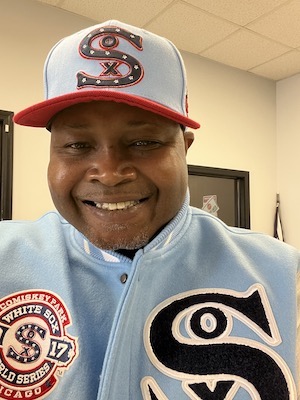 A photo of Prentice Hill wearing a red, white and blue Chicago White Sox jacket and baseball cap, facing the camera and smiling..