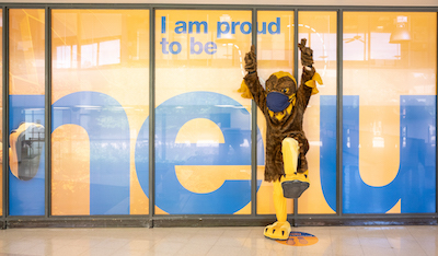 Goldie mascot in front of signage that reads "I'm proud to be NEIU"