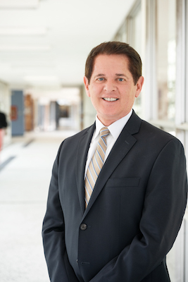 A photo portrait of R. Shayne Cofer in a black suit with white button down shirt and multicolored striped tie, smiling.