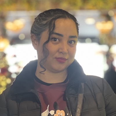 A headshot photo of Roqia Alirezaei, smiling, wearing a black jacket with yellow lights in the background.