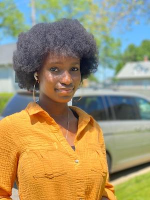 A headshot photo of Velma Elba wearing a bright yellow shirt with trees in the background.