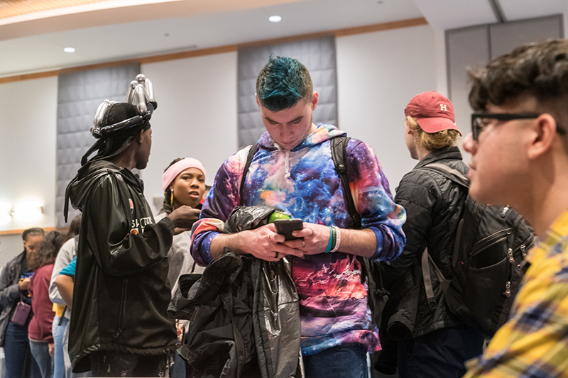 A student with blue died hair and a cosmic print shirt looks down at his phone among the crowded party.