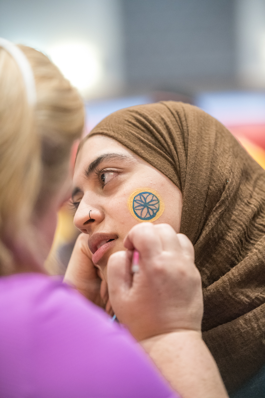 Student turns her head sideways while body artist paints a flower on her cheek in NEIU blue and gold