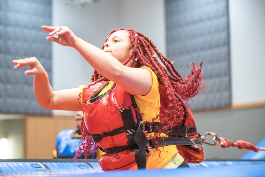 Student with bright red dreadlocks in inflatable bungee-basketball court stretches bungee chord has arms in air waiting for shot to land
