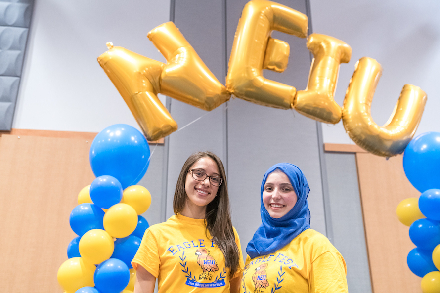 Two female students pose underneath golden Mylar balloons that spell out NEIU