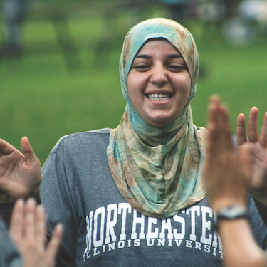 A student outdoors wearing an NEIU shirt and a headscarf smiles 