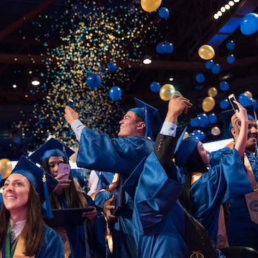 Blue and gold balloons and confetti shower graduates who are wearing regalia 