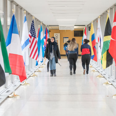 People walk past the flags of many nations line the glass hallway near the B Building