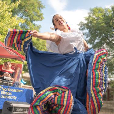 A folkloric dancer performs outdoors in a blue skirt with rainbow trim and white blouse at Northeastern Illinois University. She is smiling as she dances. 