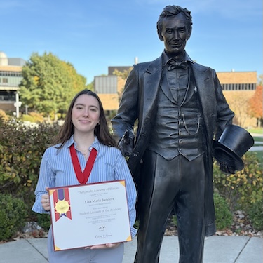 A photo of Lisa Sanders holding her Lincoln Laureate certificate and wearing her Laureate medal standing next to a statue of Abraham Lincoln. 
