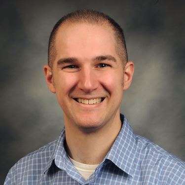 Photo of Aaron Schirmer smiling, wearing a blue and white checkered shirt against a gray background