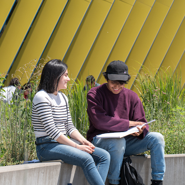 Two students sit next to each other outdoors on a low wall