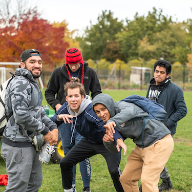 Six students hanging out together outdoors with autumnal trees in the background