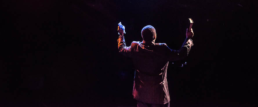A photo from a past Stage Center Theatre production; a man in a suit with arms outstretched, holding a book and a cloth tissue facing away from the camera, into a light