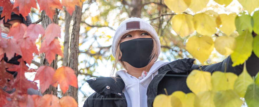 Photo of a student wearing a black coat, hooded sweater, beanie hat and black face mask surrounded by multicolored leaves with trees and branches in the background.