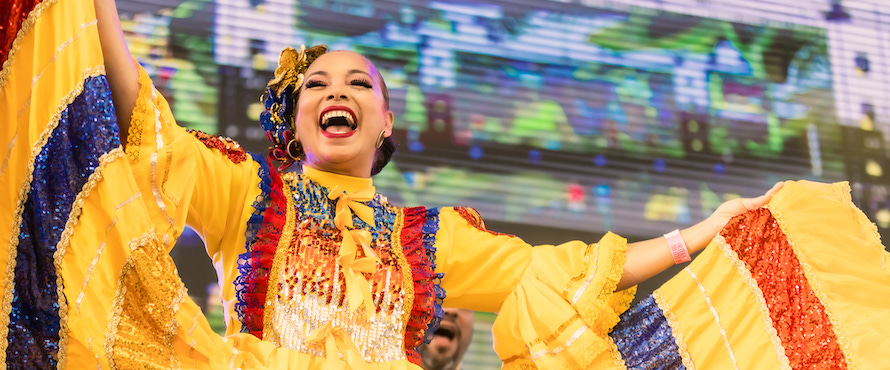 A photo of a dancer in a bright yellow, blue and red sequin folkloric dress with her holding dress ends outstretched in a dance pose, smiling, during Colombia Fest Miami 2021. Photo by Colombian Fest Worldwide.