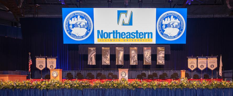 A photo of a stage with flowers along the edge, set up for Northeastern Illinois University Commencement, with two podiums, chairs, banners for the University's Values and colleges, and a screen with the Presidential Seal to the right and left of the University logo.  