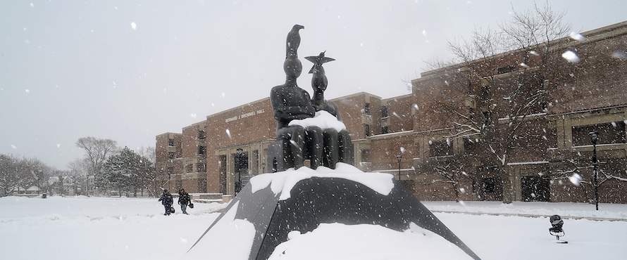 A photo of the "Serenity" statue on the Main Campus, covered in snow. Two people are walking in the background near Bernard J. Brommel Hall.
