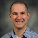 Dr. Aaron Schirmer smiles into the camera in front of a plain background.
