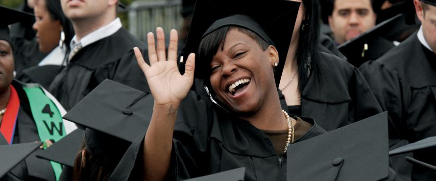 A student in a cap and gown smiles and waves to the camera.