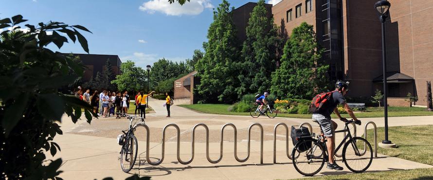 Students riding bicycles near the FA building bike rack on a sunny day