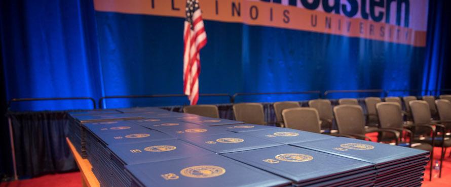 A stack of diplomas on a table with the United States flag and Northeastern Illinois University logo in the background